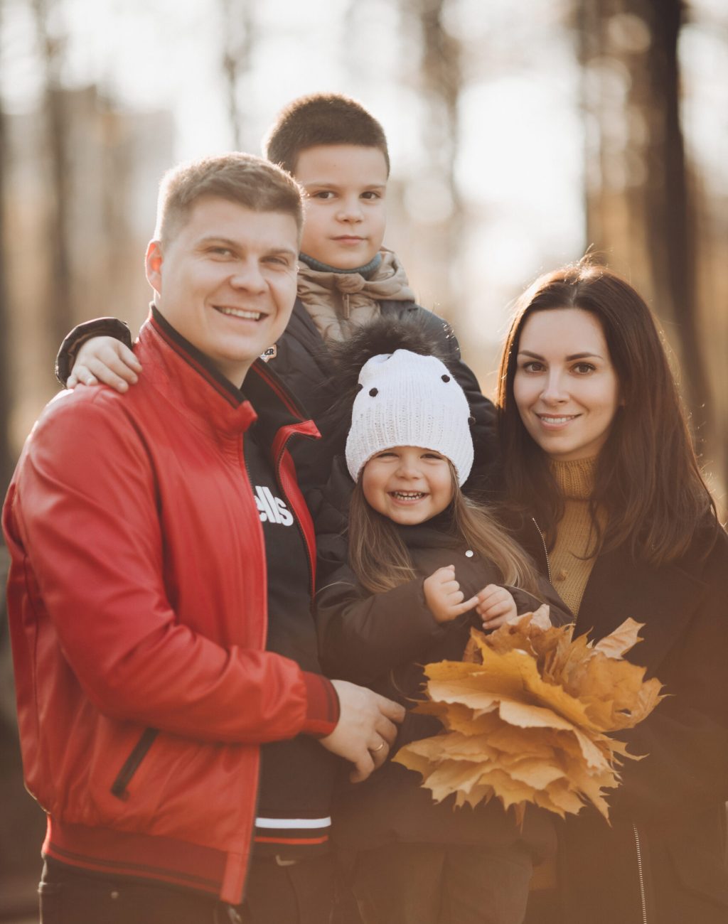 Stock photo portrait of a beautiful cheerful family with son and daughter holding bunch of yellow leaves and laughing at camera. Jolly Caucasian family together in sunny autumnal park.